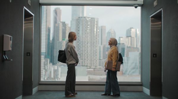 Tia stands in front of a large window with a friend. The Melbourne skyline is visible through the window behind them.