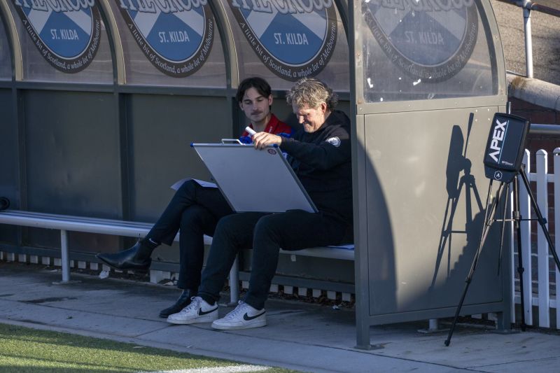 Tom and another man examine a whiteboard while sitting on a bench on the side of a sports field.