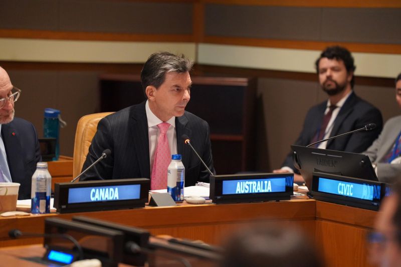 James Larsen sits in a UN committee(?) meeting with a digital sign on the desk in front of him that reads 