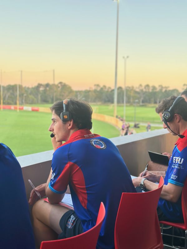 Tom sits by a sports field wearing a jersey and headset.