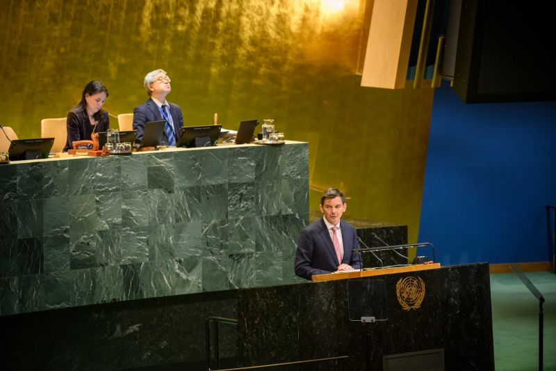 Ambassador James Larsen behind the podium at the UN General Assembly, with a wide view that shows the raised table above and behind him where people are seated.