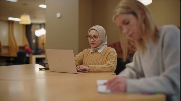 Tia works on her laptop, sitting at a table in the Melbourne Law School's Law Library with her friend.