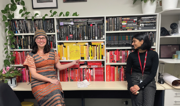 Dr Amanda Porter and Sharmera Kesavan Two people sitting on table in front of bookcase filled with books in Aboriginal flag pattern